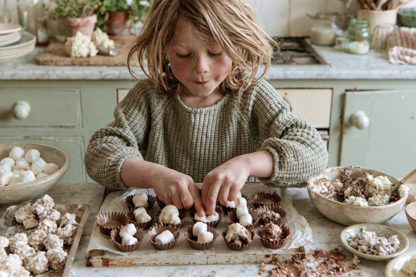 Kid pressing marshmallows into chocolate shells at a kitchen table